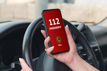 Obraz premium Woman with a manicure holds a smartphone in front of the steering wheel of a car and dialing emergency number 112. Calling emergency services on the road at the time of an accident.