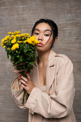 Portrait of asian model in beige trench coat holding chrysanthemums on abstract brown background