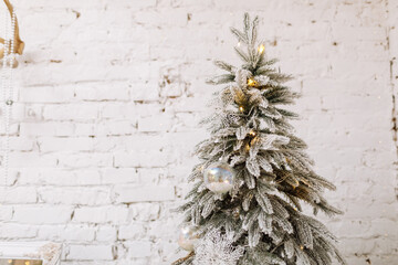 Fir tree decorated with Xmas balls on white brick wall . Close-up view of beautiful Christmas tree with white artificial snow and illuminated garland. Sparkling and fairy background. New year holiday