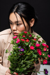 Pretty asian woman in trench coat holding colorful bouquet on black background