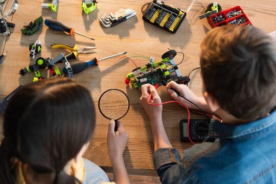 top view of boy with multimeter and girl with magnifying glass assembling robotics model at home.