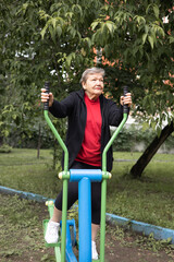 Smiling elderly woman is doing exercises on simulator outdoors in the yard.  Active life of pensioners. Adaptation of pensioners in the modern world. Prevention of Alzheimer's disease, sclerosis etc