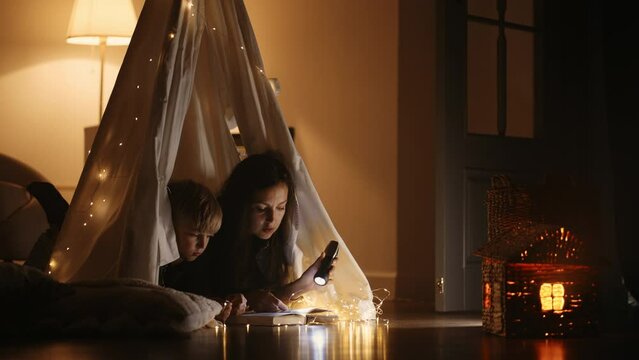 Hispanic mother and son read a book together by the light of a lantern and a tent at home in the dark by the light of garlands and lamps. Magical cozy atmosphere at home and in the family