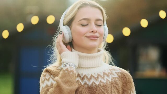 Portrait of beautiful girl relaxing while listening music in headphones in the city centre. Close up of attractive blonde woman.