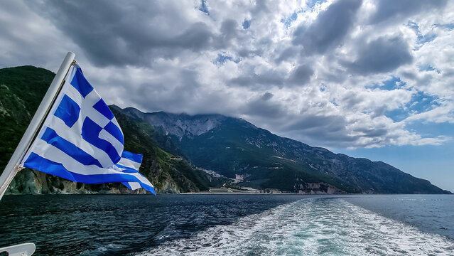 National flag of Greece waving in wind. Scenic view from tourist boat on cloud covered mountain summit Mount Athos, Chalkidiki, Central Macedonia, Greece, Europe. Greek banner in Mediterranean Sea