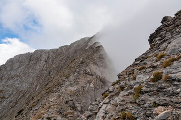 Fototapeta na wymiar Climbing on mystical foggy hiking trail leading to Mount Olympus (Mytikas, Skala, Stefani) in Mt Olympus National Park, Thessaly, Greece, Europe. Scenic view of cloud covered slopes and rocky ridges