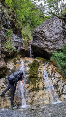 Obraz premium Man with hiking backpack drinking water from famous waterfall located in the Olympus mountains, where God Zeus used to bath. Waterfall at Mount Olympus, Thessaly, Greece, Europe. Shower of Zeus