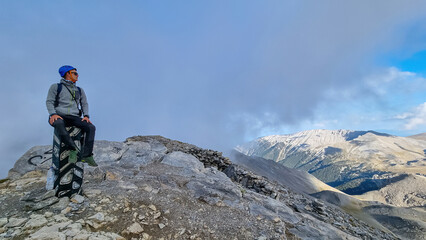 Fototapeta premium Man with helmet sitting on cloud covered mountain summit of Skolio peak on Mount Olympus, Mt Olympus National Park, Macedonia, Greece, Europe. View of rocky ridges and highlands from throne of Zeus