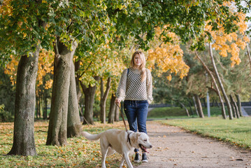 Young blonde is walking in the park with a labrador dog in the fall.