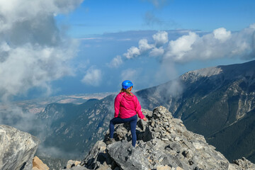 Rear view of woman with climbing helmet on cloud covered mountain summit Mytikas Mount Olympus, Mt Olympus National Park, Macedonia, Greece, Europe. View of rocky ridges and Mediterranean Aegean Sea - obrazy, fototapety, plakaty