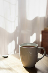 A mug full of coffee or tea set on a wooden table, morning sun and in the background light pink curtains