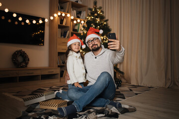 Young happy father and little cute daughter in santa hats having video call or doing selfie while sitting with smart phone in front of decorating Christmas tree in living room.