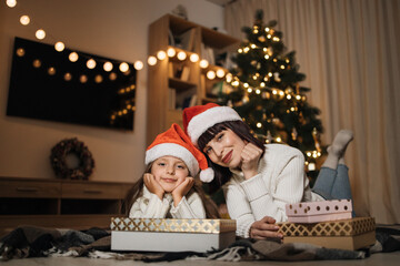 Fototapeta na wymiar Beautiful young caucasian mother lying on floor with her cute preschooler daughter with gifts in front of blurred Christmas tree in decorated room at home at evening time.