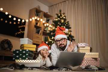 Young caucasian father and his cute little daughter chatting with somebody using laptop, lying on floor in living room at home decorated with Christmas tree.