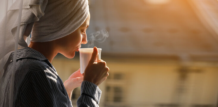 Banner of young beautiful woman is drinking hot steaming coffee or tea in sunshine light on balcony in hotel or home. Fresh girl after shower wearing in towel on head relaxing in the morning.