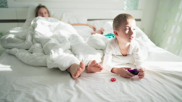 Home life brother and sister or mother in bedroom lie on bed with white linens. Sun rays in home interior in morning. Baby boy is playing with transformer robot toy. Family happy leisure activity.