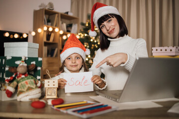 Smiling beautiful caucasian mother and her small kid girl in red hat holding letter with sale inscription on fir tree background, at evening decorated home. Concept of marry Christmas and New Year