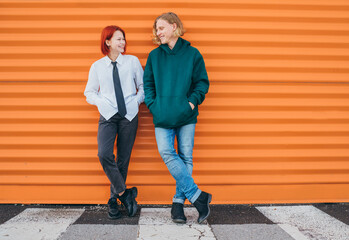 Portraits of two smiling to each other caucasian teen friends boy and girl casual dressed while they standing lean on the orange wall background. Careless young teenhood time and first love concept.