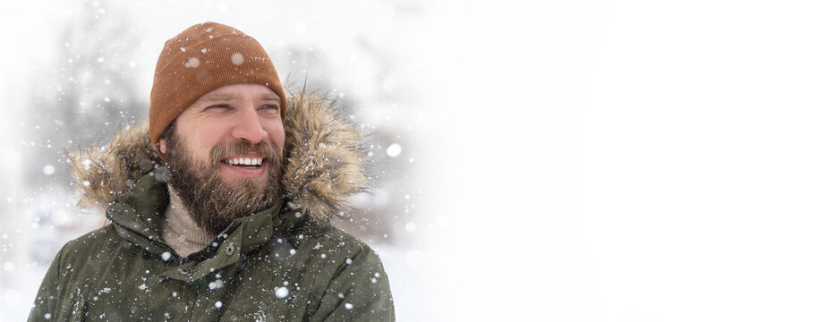 Close up mature caucasian man bearded winter coat with fur collar and brown knitted hat snow outdoor. Positive toothy smile looking to the side. Mock up, copy space extra wide banner.