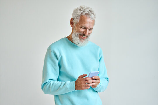 Older Happy Senior Bearded Stylish Hipster Man Holding Cell Phone Looking At Cellphone Using Smartphone Checking Mobile Apps News Or Communicating Online Standing Isolated On White Wall.