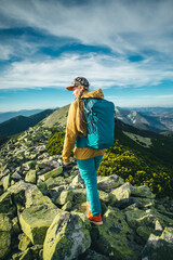 a girl hiker in the mountains with a blue backpack walks against the background of incredibly beautiful mountains. A girl in a cap and a mustard-colored jacket walks along the rocks