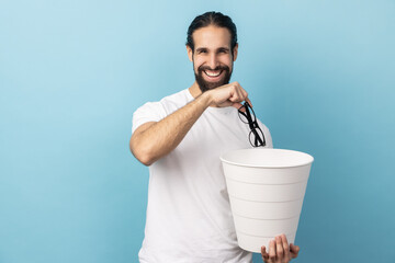 Portrait of man with beard wearing white T-shirt throwing out eyeglasses into rubbish bin, treat his eyesight, looking smiling at camera. Indoor studio shot isolated on blue background.