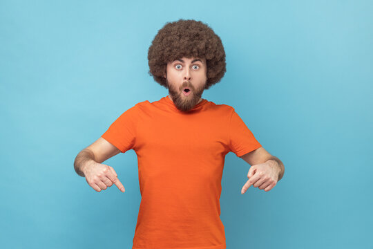 Portrait of shocked surprised man with Afro hairstyle wearing orange T-shirt pointing down with fore fingers, looking at camera with open mouth. Indoor studio shot isolated on blue background.
