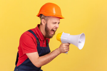 Side view of angry bearded worker wearing protective helmet and blue overalls holding megaphone and...