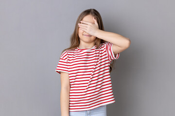 Portrait of dark haired little girl wearing striped T-shirt standing, covering eyes with hand, refusing to watch, shame content. Indoor studio shot isolated on gray background.