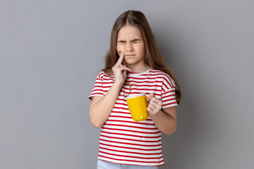 Portrait of unhealthy little girl wearing striped T-shirt touching chin suffering tooth ache, has...