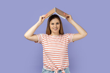 Portrait of beautiful attractive woman wearing striped T-shirt holding opened book on head, looking...