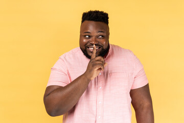 Portrait of optimistic positive man wearing pink shirt standing, showing hush sign and looking away with toothy smile, sharing secret. Indoor studio shot isolated on yellow background.