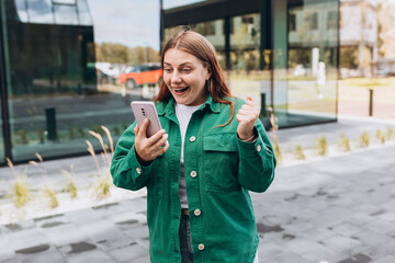 Fototapeta premium Portrait of a satisfied young woman holding mobile phone and celebrating outdoors. Happy girl walking on city street, hipster female overjoyed with winning online contest on website