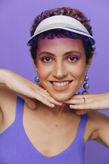 Portrait of a sporty fashion woman posing smiling with teeth at the camera with stylish earrings in her ears in a purple yoga tracksuit and a transparent cap on a purple monochrome background