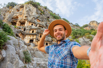 Swarthy caucasian european guy in plaid shirt smiles rejoices makes a selfie in a brown hat at the ancient unique landmarks Turkey, Antalya Demre, Myra, ancient lycian tomb cave ruins.
