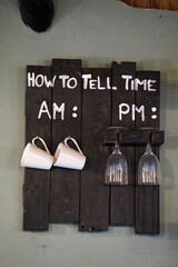 Wooden board mounted on wall with a funny joke of how to tell time between glasses and cups with soft side lighting. Mounted inside a bar that serves breakfast