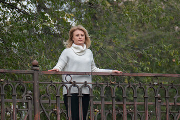Serious dreamy European lady concentrated somewhere. Middle-aged blonde woman on a walk in the autumn park. Pensive woman in white sweater stands on the city bridge and enjoys nature.