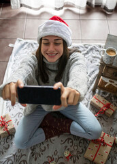 Christmas vacations online. Happy woman having a video call with her family or friends. Young woman using a mobile phone taking a selfie sitting on the floor on a blanket with gifts. Virtual meeting