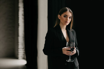Portrait of successful woman in formal jacket, holding a glass of wine and looking at camera. Serious woman, with professional makeup, dressed in black suit, standing in empty dark hall of building