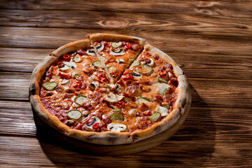Appetizing pizza with hunting sausages, mozzarella cheese, pickled cucumbers, chili peppers on a tomato base on wood background