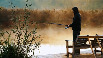 Fisherman throwing fishing rod at the background autumn landscape morning haze shrouded river.
