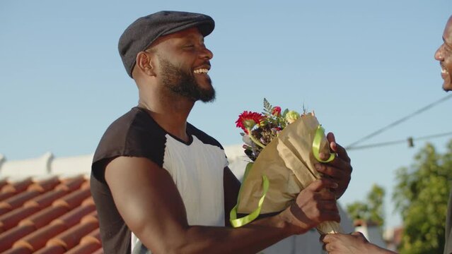 Medium shot of happy bearded gay taking flowers from lover. Side view of embarrassed Afro-American men having first date on rooftop, hugging and kissing cheek, showing affection. LGBT, love concept