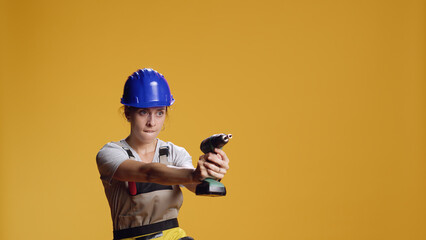 Construction worker having fun with power drill machine, fooling around in studio and playing with electric drilling nail gun over yellow background. Playful female handywoman with screw gun.