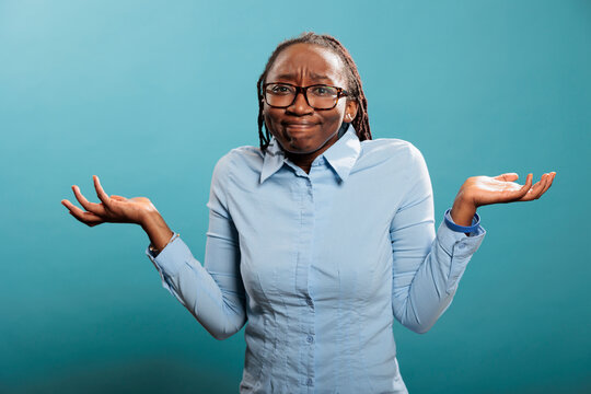 Puzzled and doubtful young adult person being hesitant and unconfident while answering question. Confused and uncertain woman shrugging shoulders with uncertainty while standing on blue background.