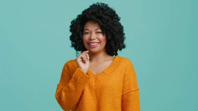Joyful dark skinned woman feels very happy points finger picking you at camera and positively winks.African American model openly smiling dressed in orange sweater isolated over blue studio background