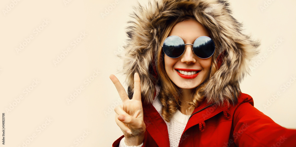 Wall mural Portrait of happy smiling woman stretching hand for taking selfie with smartphone wearing jacket with fur hood