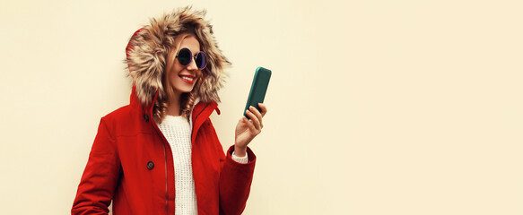 Portrait of stylish smiling young woman with smartphone wearing red jacket with fur hood, backpack on background, blank copy space for advertising text