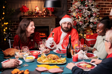 Man in santa claus costume wishing merry christmas at festive table, proposing toast, holding sparkling wine glass. Winter holidays celebration with family at home feast, diverse friends gathering