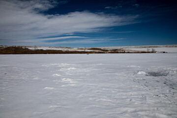 Pristine white snow covered frozen lake with ice fisherman in the distance and an abandoned fishing hole with tracks in the snow and a blue sky with white cirrus clouds
