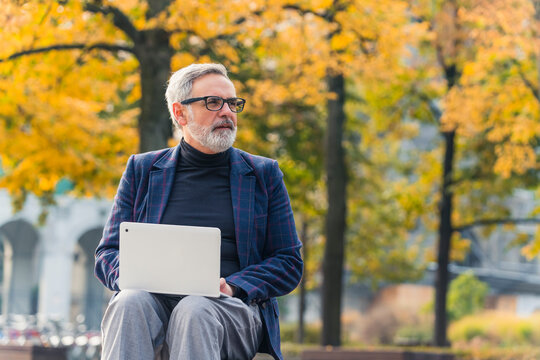 Middle-aged man with grey hair and beard wearing glasses and smart-casual clothes sitting on bench in city park working on laptop computer looking around. Horizontal outdoor shot. High quality photo